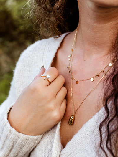 Close-up of woman wearing gold layered necklaces with teardrop pendant and disc chain, paired with pearl ring and cream knit sweater, elegant minimalist jewelry bundle for modern style.