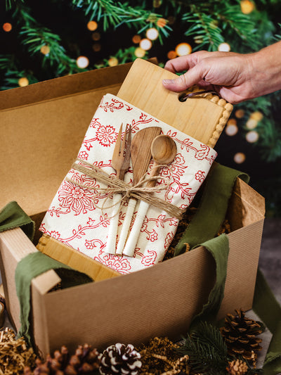 Handcrafted holiday gift box with wooden cutting board, red floral cotton tea towel, and artisan kitchen utensils tied with twine – sustainable Christmas gift idea