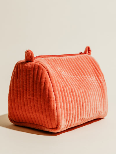 A coral pink quilted velvet cosmetic pouch with a ribbed texture, standing upright on a neutral background. Its soft design and zipper closure add functionality to its vibrant look.