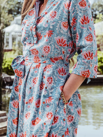 lose-up of the waist and bodice of a teal floral dress featuring coral and white flowers, a ruffled neckline, button details, and a self-tie belt. The woman's hand rests in her pocket, showing a gold bracelet and chain link jewelry.
