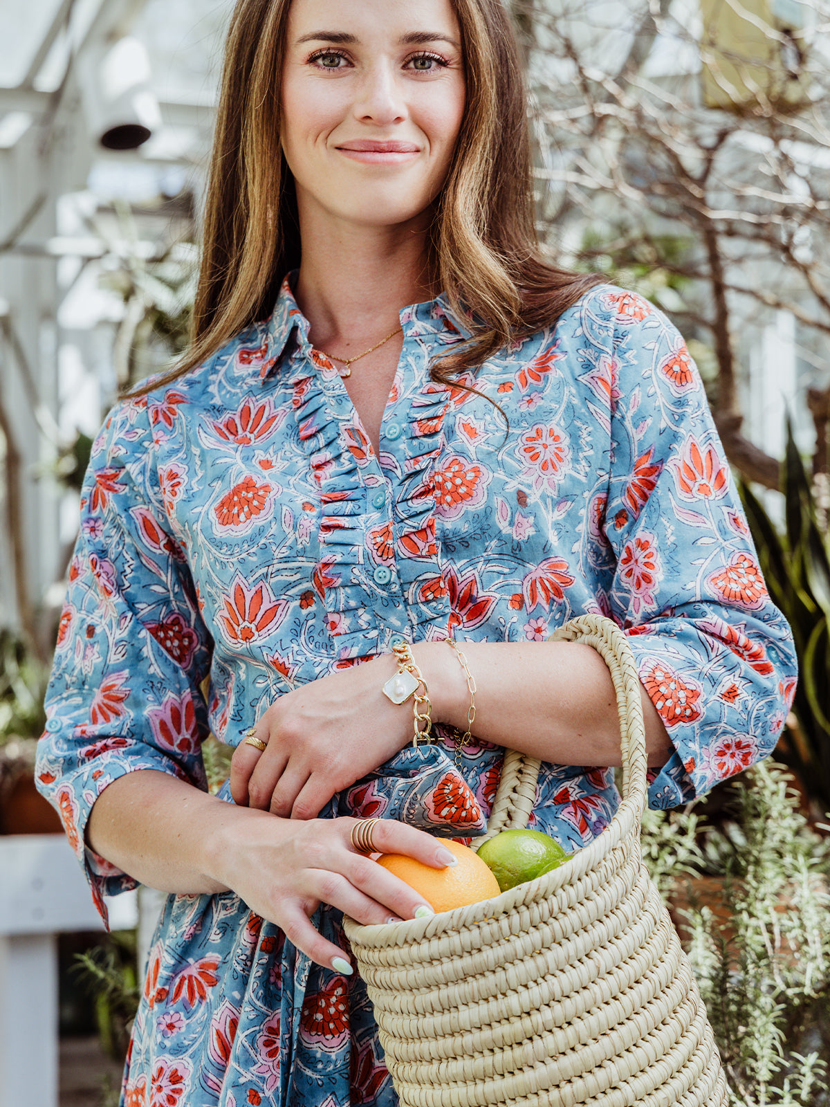 Woman smiling in a teal floral print dress with a ruffled neckline, holding a woven basket of citrus. The setting is a greenhouse, and she wears gold bracelets, rings, and delicate chain jewelry.
