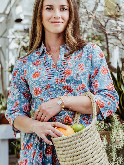 Woman smiling in a teal floral print dress with a ruffled neckline, holding a woven basket of citrus. The setting is a greenhouse, and she wears gold bracelets, rings, and delicate chain jewelry.