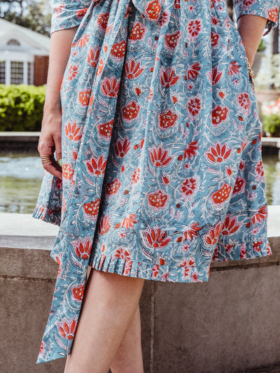 Close-up of the lower half of a teal floral wrap dress with coral flowers, highlighting the fabric’s texture, pleated hemline, and tie waist detail. The woman’s hand rests casually by her side, with gold rings and a visible bracelet.