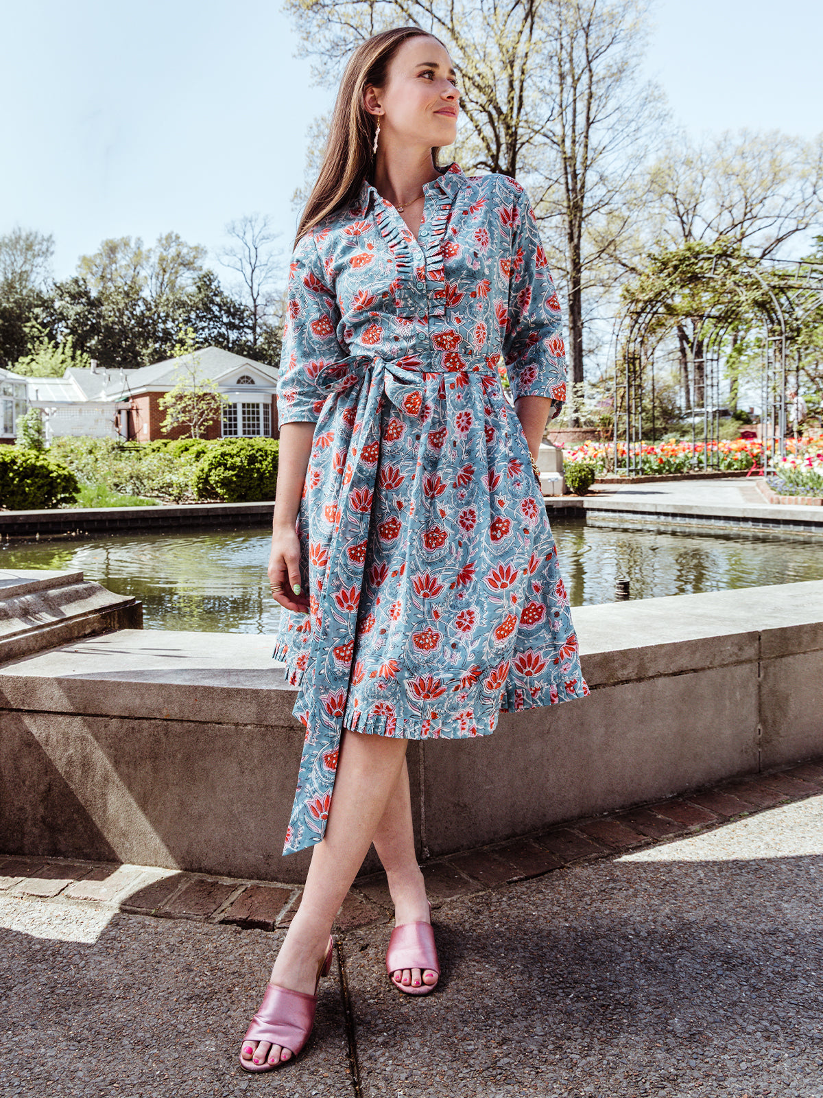 Full-body view of a woman in a floral midi wrap dress with a ruffled neckline and tie waist, styled with pink satin heeled sandals. She stands in front of a pond and blooming garden on a sunny spring day.