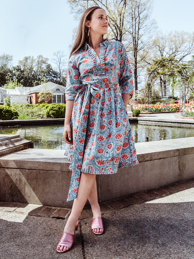 Full-body view of a woman in a floral midi wrap dress with a ruffled neckline and tie waist, styled with pink satin heeled sandals. She stands in front of a pond and blooming garden on a sunny spring day.
