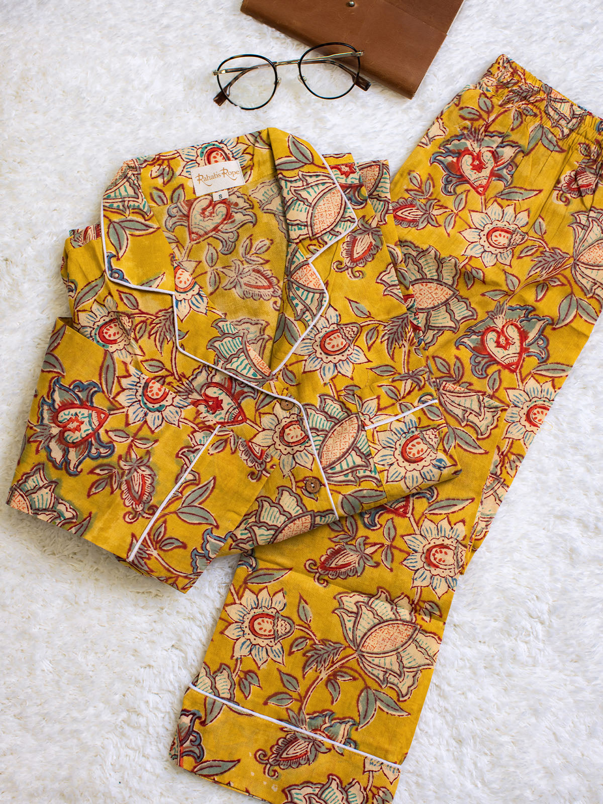 Folded yellow floral pajama set with white piping, displayed on a soft white surface beside round eyeglasses and a brown leather notebook.