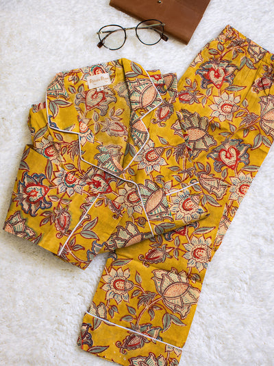 Folded yellow floral pajama set with white piping, displayed on a soft white surface beside round eyeglasses and a brown leather notebook.