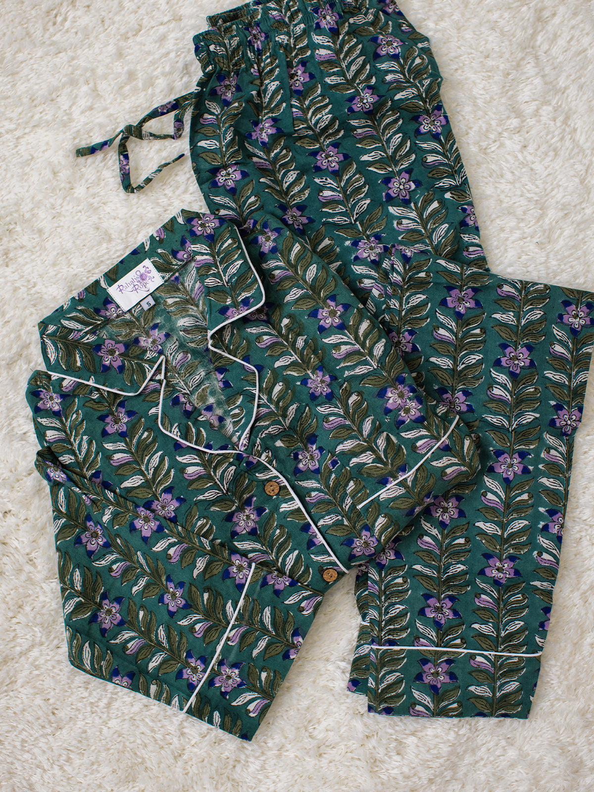 Folded teal pajama set with purple and olive green floral patterns and white piping, featuring a drawstring waistband, laid on a soft cream rug.