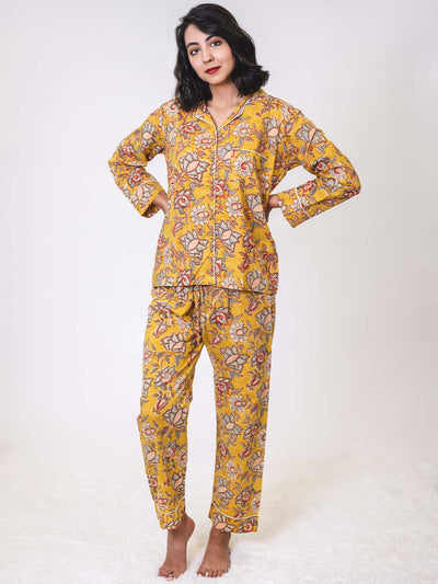 Woman standing barefoot on a white rug, wearing a yellow floral two-piece pajama set with long sleeves and full-length pants, hands resting on her hips, looking confidently at the camera.