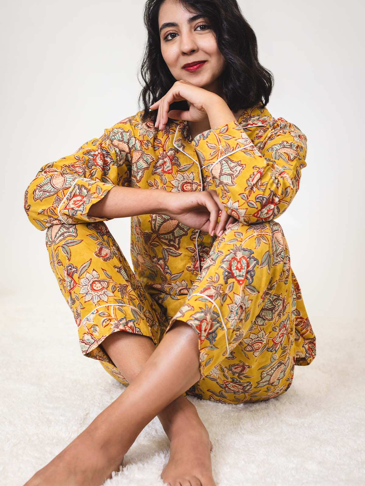 Woman sitting cross-legged on a white rug, wearing a yellow floral pajama set, resting her chin on her hand and smiling softly at the camera.
