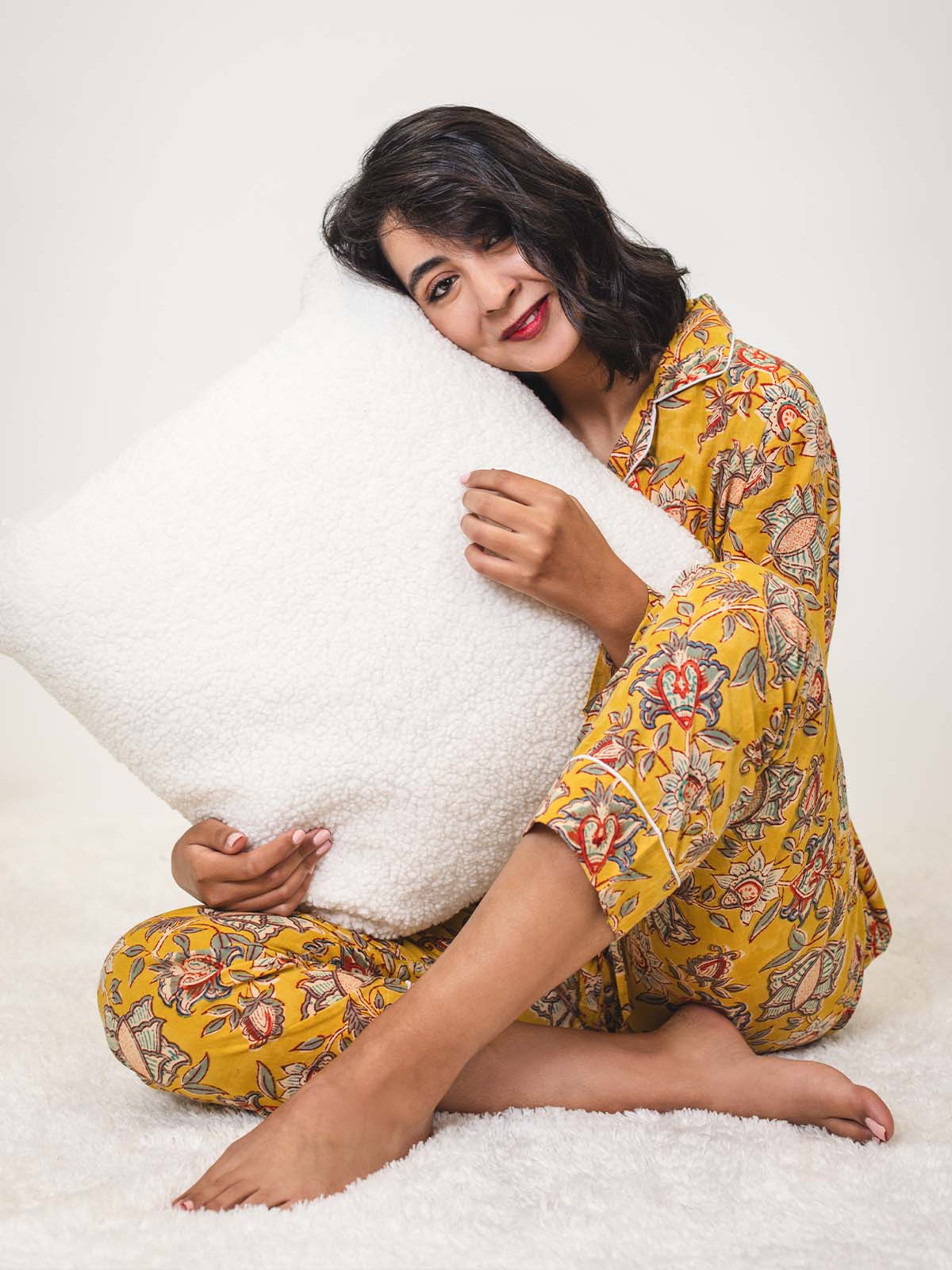 Woman sitting cross-legged on a white rug in yellow floral pajamas, hugging a large white textured pillow and smiling warmly.