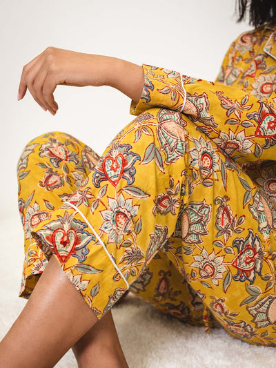 Close-up of a woman’s legs and arm while sitting cross-legged in yellow floral pajamas, highlighting the intricate floral pattern and white piping at the cuffs.