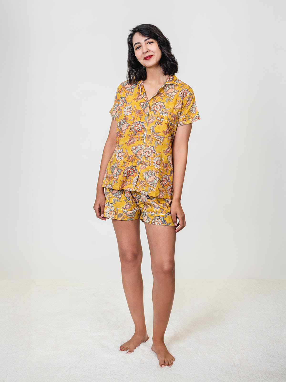 Woman standing barefoot on a white rug, wearing a yellow floral short-sleeve pajama set with shorts, smiling softly at the camera.