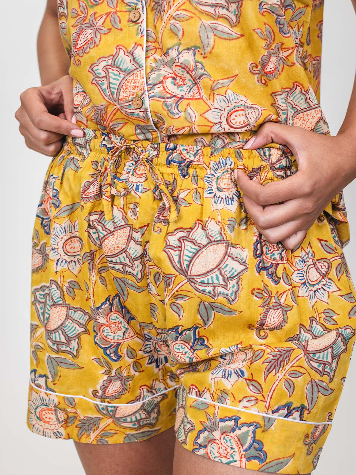 Close-up of a woman wearing yellow floral pajama shorts, holding the elastic waistband with both hands to show the drawstring detail.