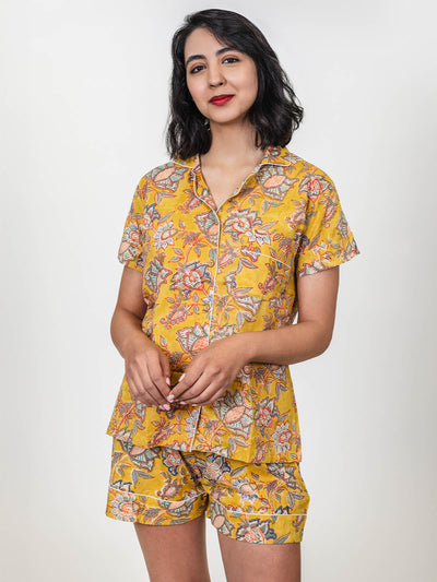  Woman in a yellow floral short-sleeve pajama set with shorts, standing and smiling slightly, hands clasped in front of her.