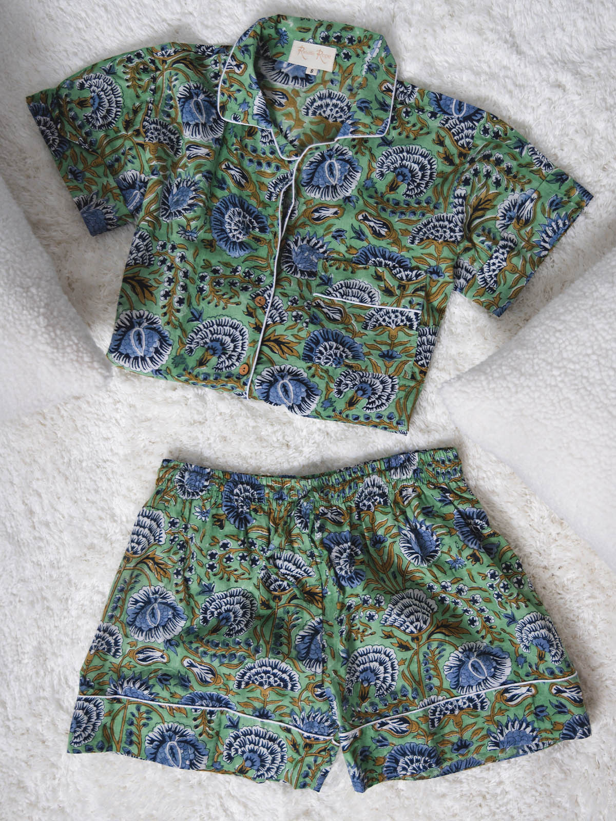 Folded green short-sleeve pajama top and matching shorts with blue and white floral print and white piping, displayed on a soft white textured surface.