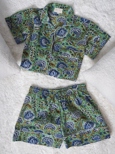Folded green short-sleeve pajama top and matching shorts with blue and white floral print and white piping, displayed on a soft white textured surface.