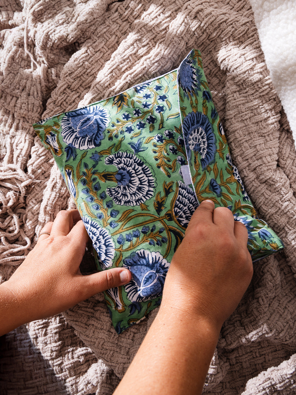Hands holding a handmade fabric pouch with a green background and bold blue floral block print design, placed on a soft textured beige blanket.