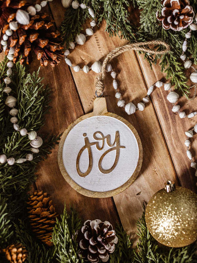 Rustic wooden Christmas ornament with the word 'joy' carved on white background, styled with pinecones, evergreen garland, and gold bauble holiday decor.