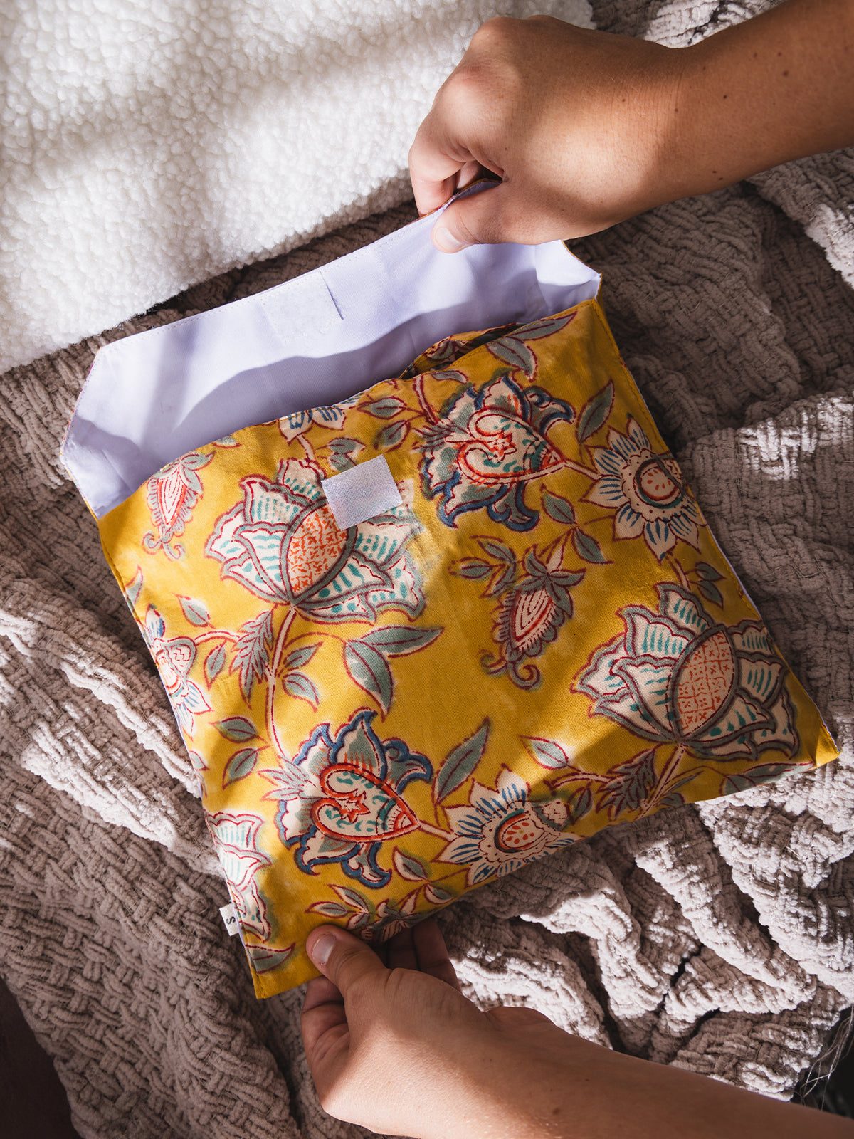 Hands opening a handmade fabric pouch with a bright yellow background and red-blue floral block print design, set on a cozy beige textured blanket.