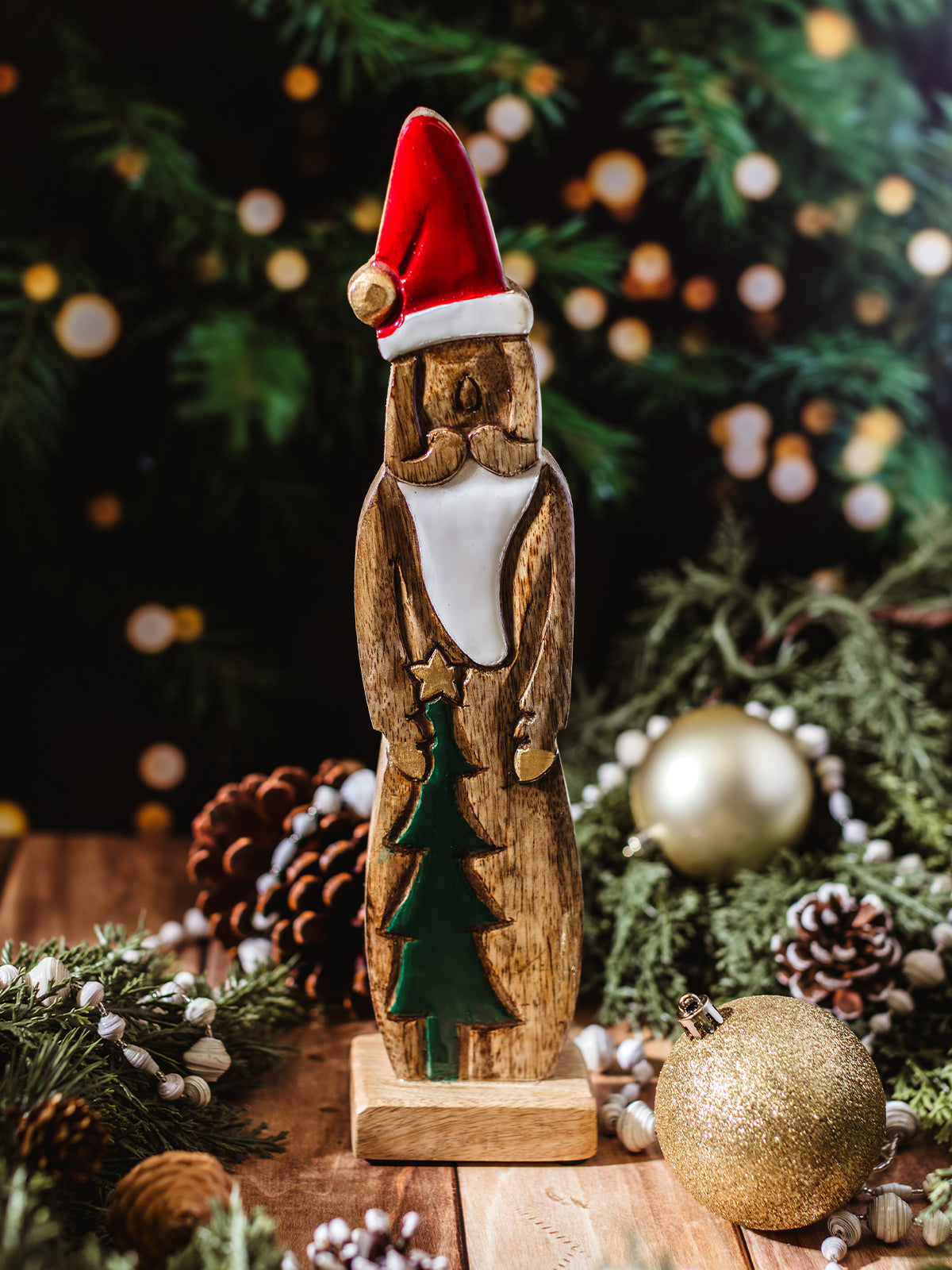 Carved wooden Santa Claus figurine with red hat, white beard, and green Christmas tree detail, surrounded by pinecones, ornaments, and holiday greenery.