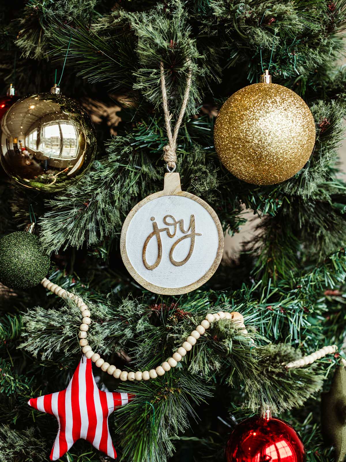 Round wooden “joy” ornament with rustic twine hanger displayed on a Christmas tree decorated with gold, red, and green baubles, wooden bead garland, and a red-striped star — handmade holiday decoration.
