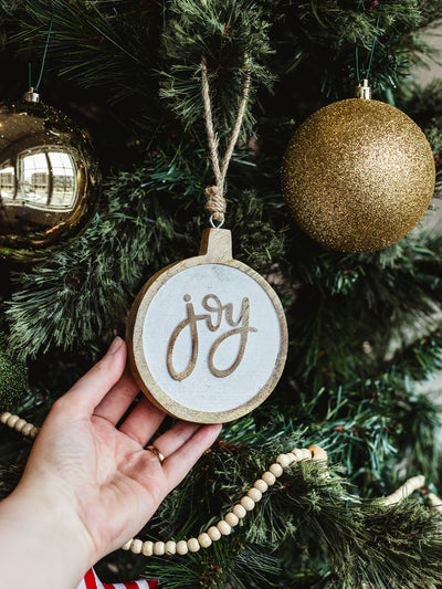 Hand holding a round wooden “joy” Christmas ornament with rustic twine hanger on a decorated holiday tree with gold baubles and wooden bead garland — handcrafted ethical décor.