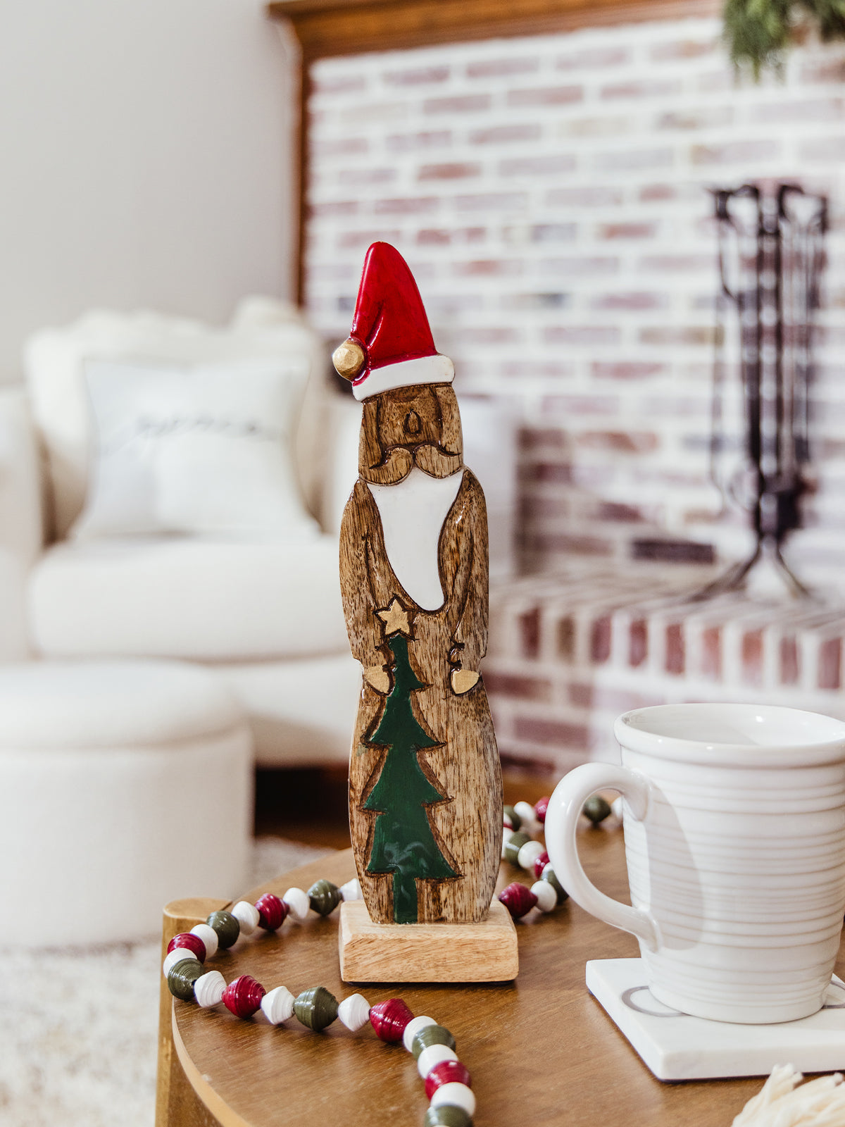 Handcrafted wooden Santa figurine with red hat and green Christmas tree detail displayed on a cozy coffee table with holiday bead garland and a mug — rustic ethical Christmas décor.
