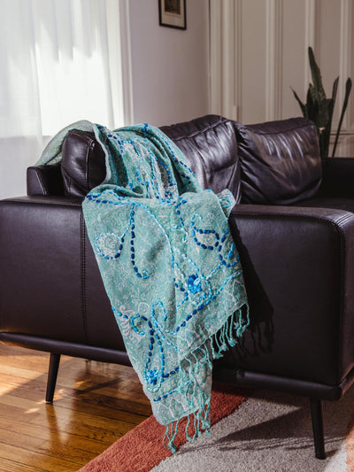 Green embroidered shawl with blue accents and fringes, elegantly draped over a black leather sofa, part of Joffa.com's artisanal home and fashion collection.