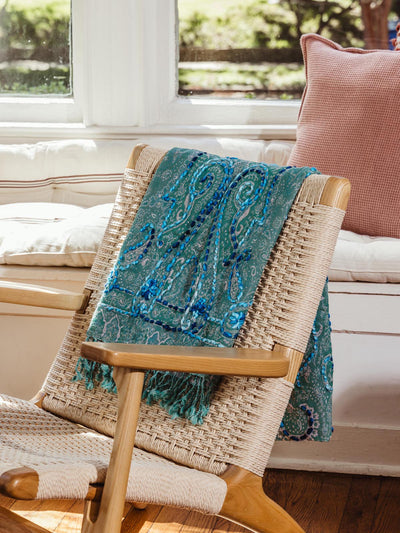 Green paisley shawl with blue embroidery and fringes draped over a woven chair, highlighting its elegant texture and vibrant colors, available at Joffa.com.