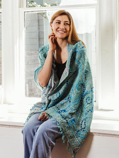 Smiling woman sitting by a window, wearing a green shawl with blue embroidered detailing, part of Joffa.com's handmade and sustainable fashion collection.