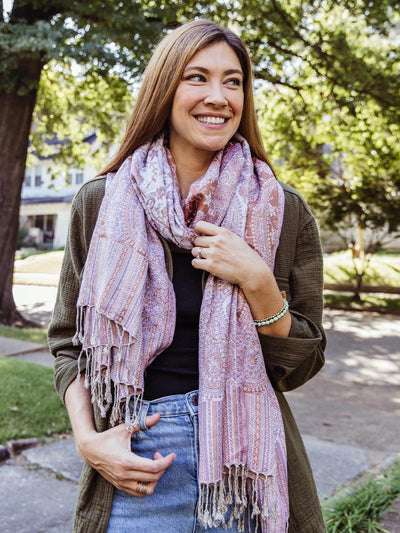 Woman wearing a pink, rust orange, and white paisley shawl with fringes, smiling outdoors, part of Joffa.com's sustainable and stylish accessory collection.
