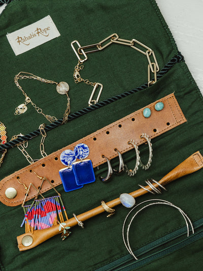 Open jewelry roll with leather strap and green fabric interior, displaying earrings and necklaces. Handcrafted by Rahab's Rope, a unique and ethical jewelry organizer available at Joffa.com.