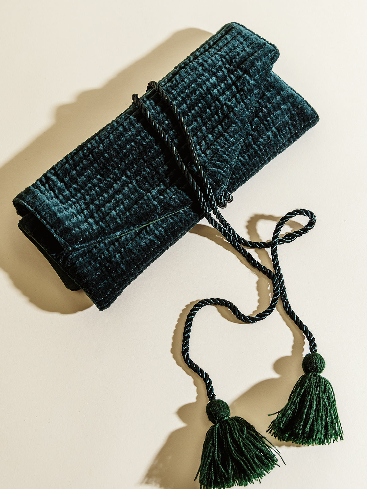 A dark green velvet jewelry roll, neatly tied with twisted rope featuring two tassels, resting on a cream background. The soft texture and elegant design highlight its luxurious, handmade appearance.