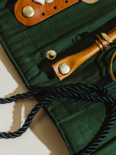  A close-up of the interior of the jewelry roll, showing a rich green fabric lining with tan leather sections and brass details for organizing jewelry. The twisted green rope and metal rings are also visible.