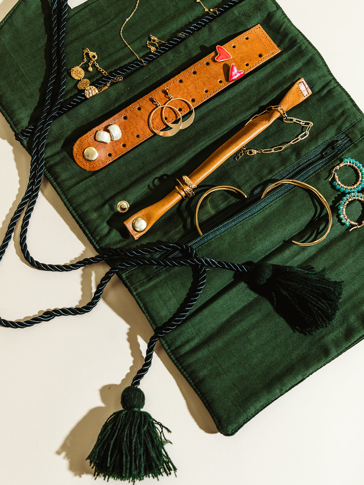 An open dark green velvet jewelry roll displaying necklaces, rings, bracelets, and earrings organized with leather straps and compartments. Twisted rope with tassels drapes elegantly over the piece.