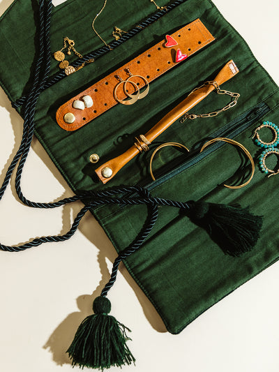 An open dark green velvet jewelry roll displaying necklaces, rings, bracelets, and earrings organized with leather straps and compartments. Twisted rope with tassels drapes elegantly over the piece.