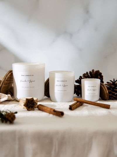 Three white glass candles from Riau Candle Co. in Vanilla & Spice scent, displayed in small, medium, and large sizes on a linen cloth with pinecones, wood slices, and cinnamon sticks for a cozy holiday feel.