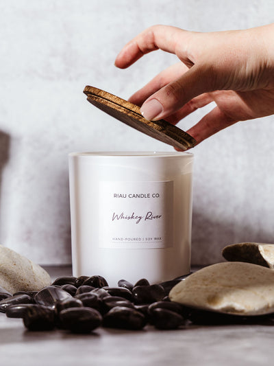 A hand gently lifts the wooden lid from a Riau Candle Co. Whiskey River small candle, styled among smooth black stones.