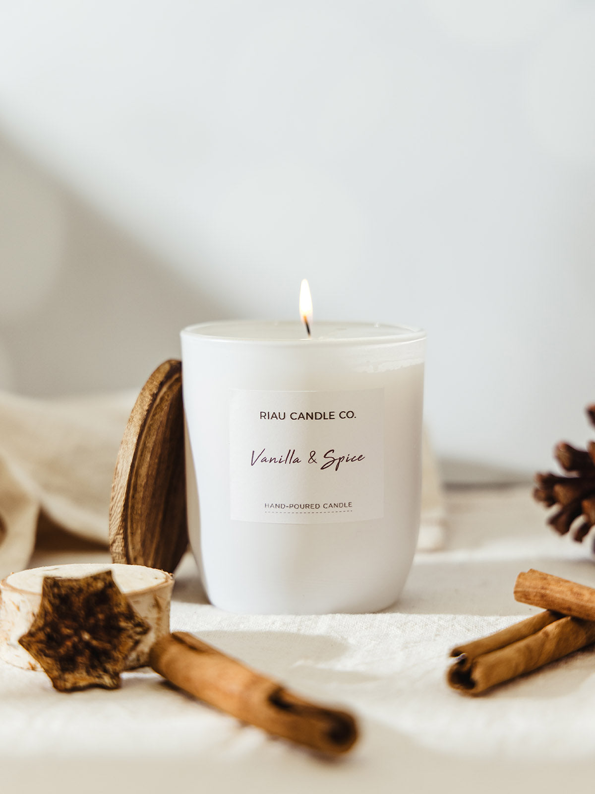 Minimalist white mini Vanilla & Spice candle by Riau Candle Co. burning softly, surrounded by cinnamon sticks and natural wood accents on a cream cloth.

