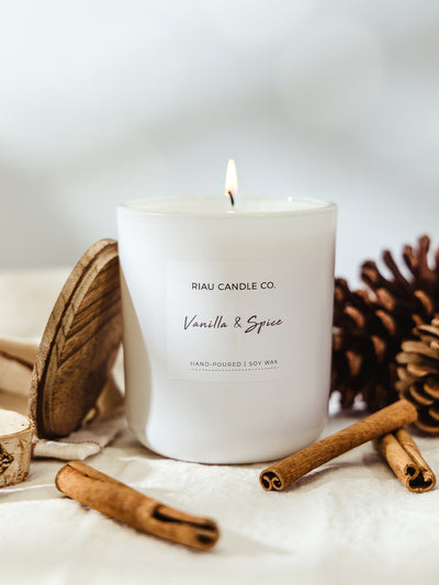 Hand-poured large Vanilla & Spice soy candle in a white glass jar from Riau Candle Co., glowing with a warm flame beside a pinecone and cinnamon sticks.