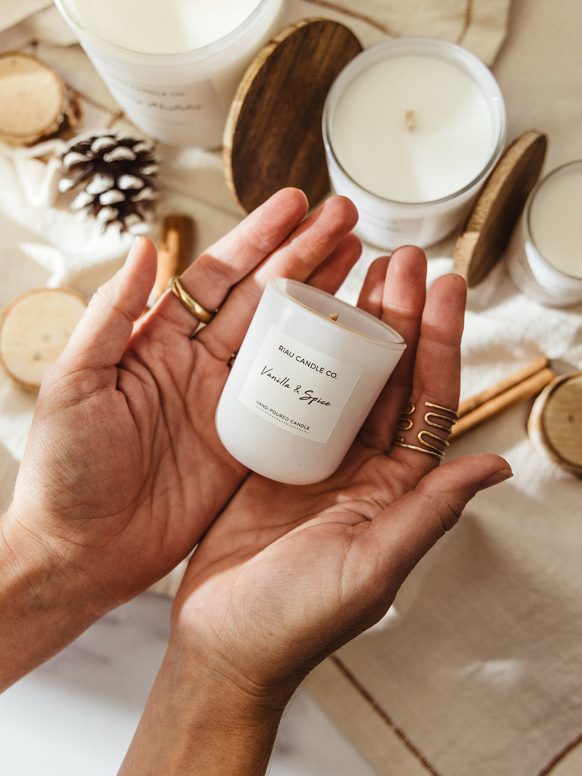 Pair of hands holding a small white Vanilla & Spice candle by Riau Candle Co., styled on a neutral linen cloth with larger candles, pinecones, wood slices, and cinnamon sticks in the background.