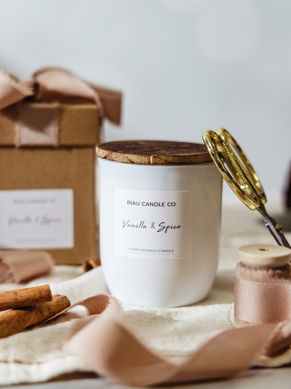 Riau Candle Co. Vanilla & Spice hand-poured soy candle in a white glass jar with wooden lid, styled with blush ribbon, cinnamon sticks, gold scissors, and a kraft gift box for a cozy holiday gift look.
