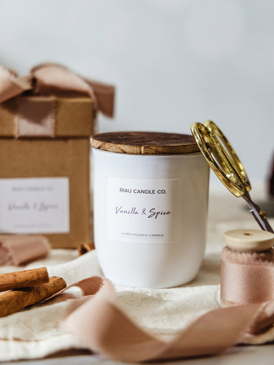 Riau Candle Co. Vanilla & Spice hand-poured soy candle in a white glass jar with wooden lid, styled with blush ribbon, cinnamon sticks, gold scissors, and a kraft gift box for a cozy holiday gift look.