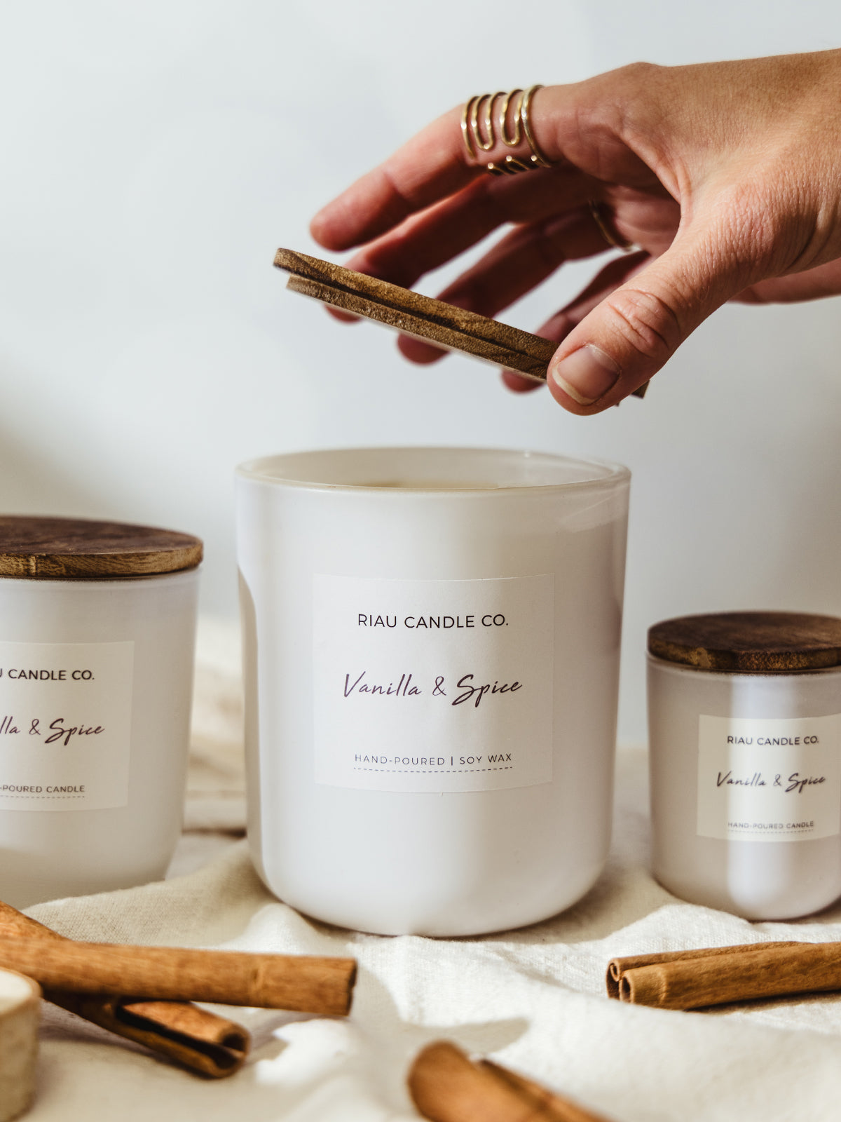 Close-up of a hand with gold rings placing a wooden lid on a large Vanilla & Spice soy candle by Riau Candle Co., surrounded by cinnamon sticks and matching candles in various sizes.