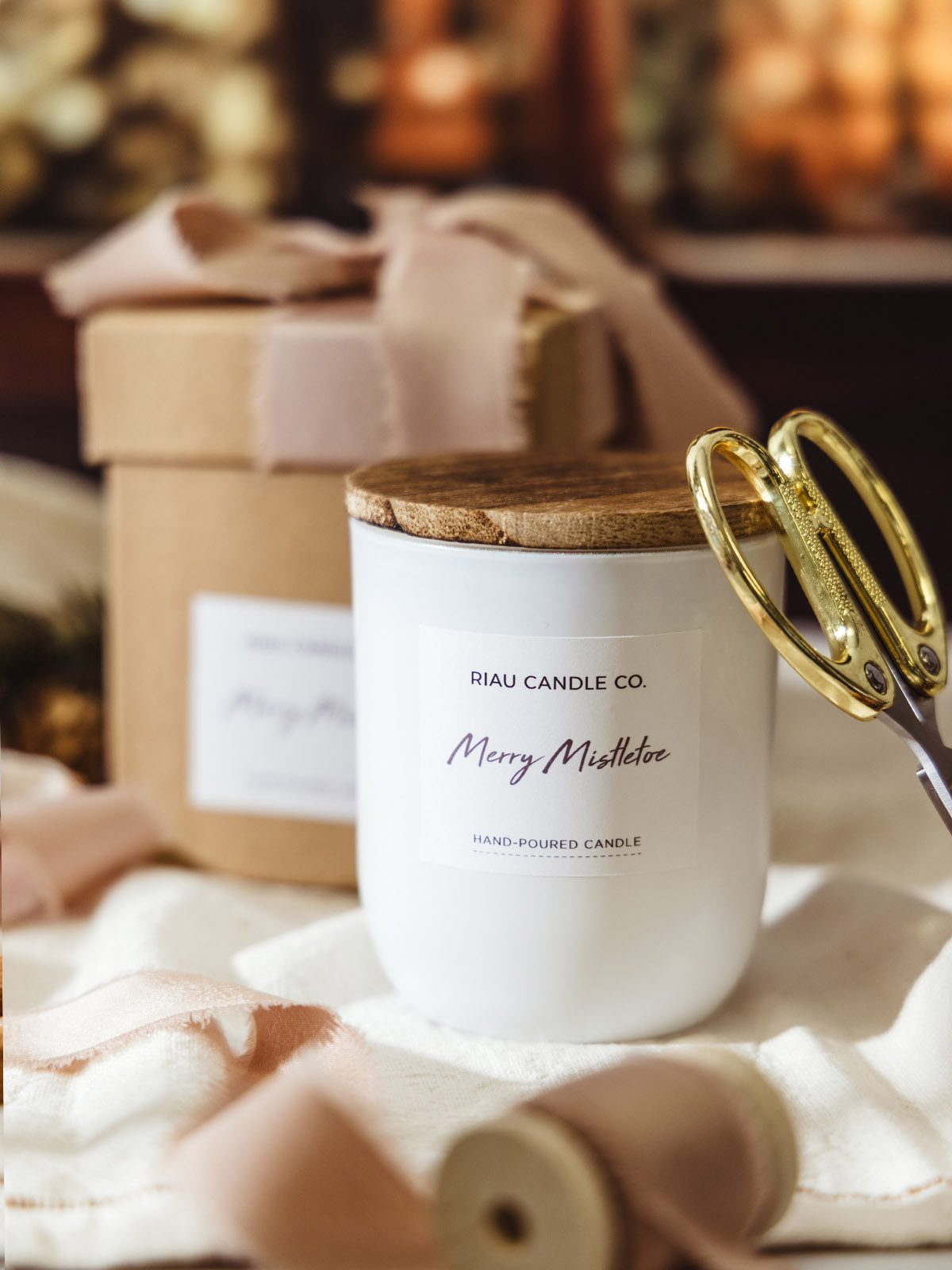 Riau Candle Co. Merry Mistletoe hand-poured soy candle in a white glass jar with wooden lid, surrounded by blush ribbon, gold scissors, and a wrapped kraft gift box, styled for festive gifting.