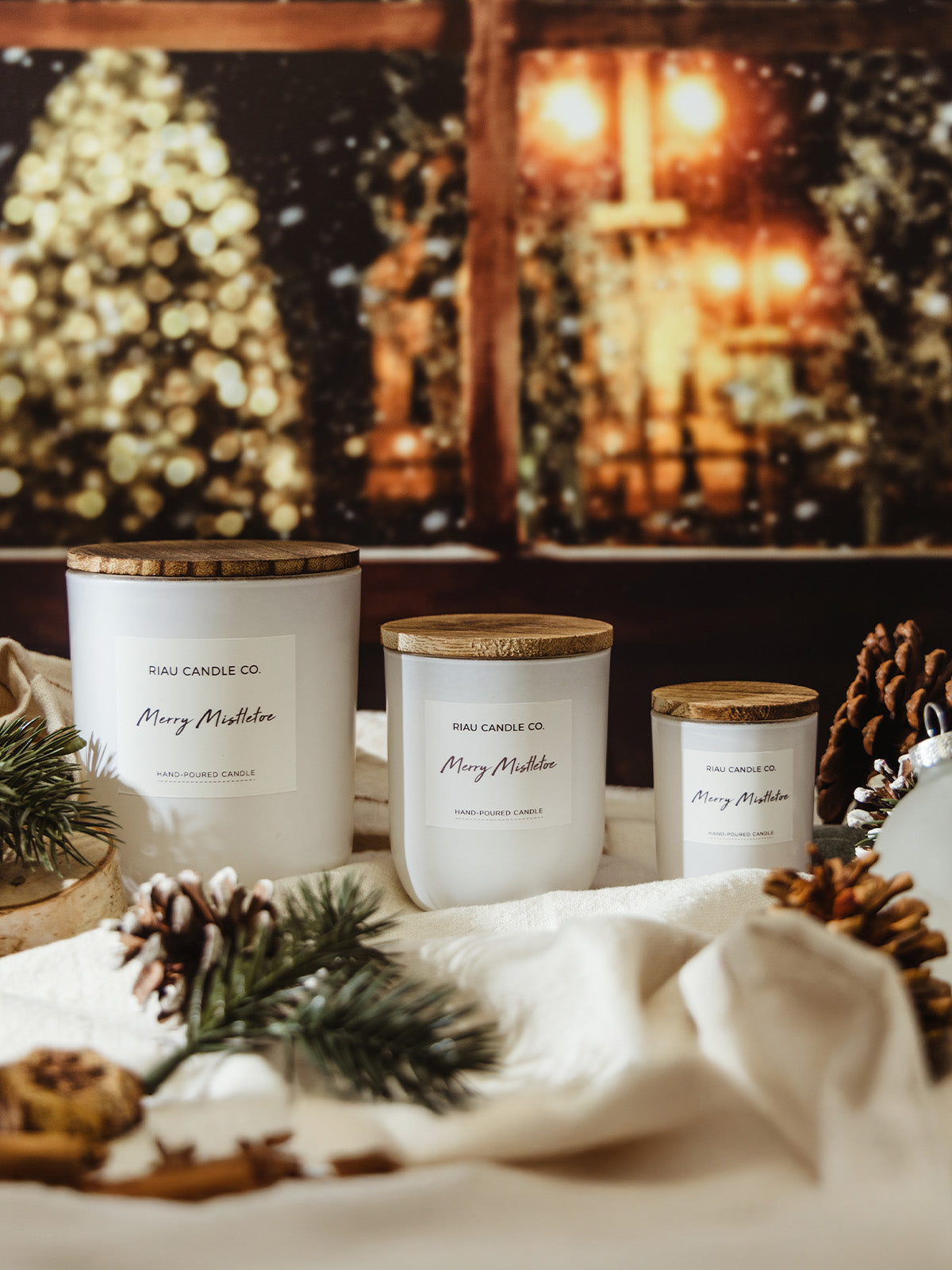 Set of three Riau Candle Co. Merry Mistletoe candles in white jars with wooden lids, styled on a cream cloth with pinecones and evergreens, against a cozy holiday background of glowing Christmas lights.
