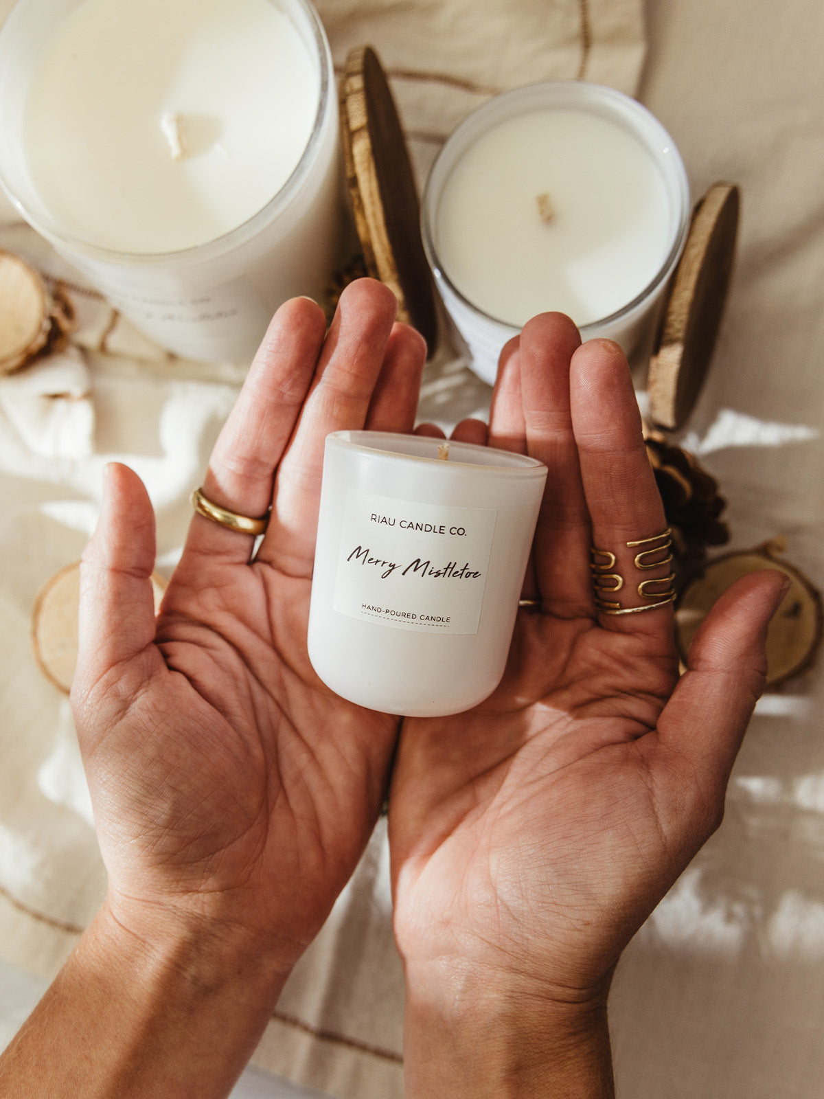 Hands holding a small Merry Mistletoe hand-poured soy candle by Riau Candle Co., with larger candles, wooden lids, and pinecones softly blurred in the background.