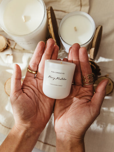 Hands holding a small Merry Mistletoe hand-poured soy candle by Riau Candle Co., with larger candles, wooden lids, and pinecones softly blurred in the background.