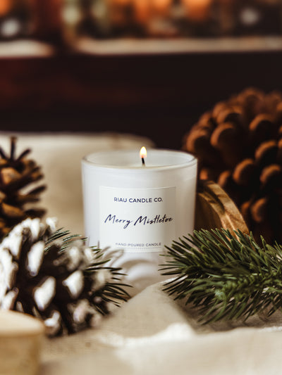 Lit Merry Mistletoe candle in a white glass jar by Riau Candle Co., surrounded by frosted pinecones and evergreen sprigs for a festive winter look.